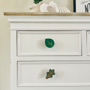 May include: White dresser with two drawers featuring green leaf-shaped drawer pulls with gold accents. The top drawer pull is a simple leaf shape, while the bottom drawer pull is an oak leaf shape.