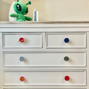 May include: White chest of drawers with four drawers and colourful planet-shaped knobs. The top drawer has a red knob, the second drawer has a blue knob, the third drawer has a grey knob, and the bottom drawer has a pink knob.