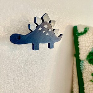 Peut inclure: Un crochet mural en forme de dinosaure imprim&eacute; en 3D de couleur bleue avec un dos &agrave; pointes blanches et des pois blancs.