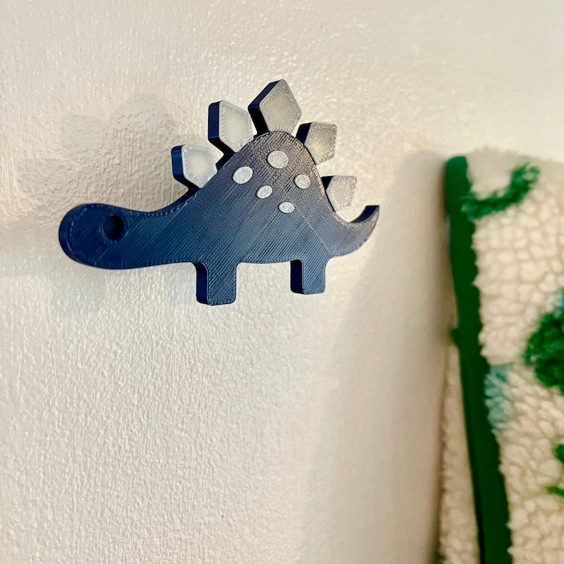 Peut inclure: Un crochet mural en forme de dinosaure imprim&eacute; en 3D de couleur bleue avec un dos &agrave; pointes blanches et des pois blancs.