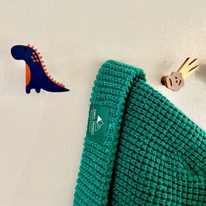 Peut inclure: Un bonnet en tricot vert avec une &eacute;tiquette blanche indiquant "Earth Friendly" et un petit logo de montagne. Le bonnet est accroch&eacute; &agrave; un mur blanc avec un dinosaure en bois et une d&eacute;coration en nuage en bois.