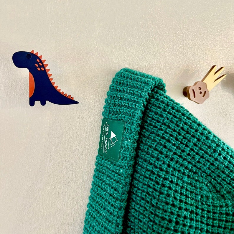 Peut inclure: Un bonnet en tricot vert avec une &eacute;tiquette blanche indiquant "Earth Friendly" et un petit logo de montagne. Le bonnet est accroch&eacute; &agrave; un mur blanc avec un dinosaure en bois et une d&eacute;coration en nuage en bois.