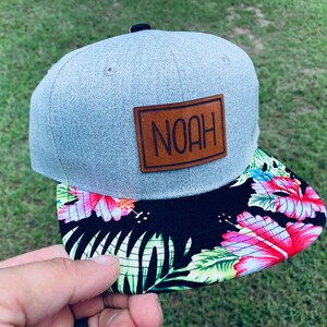 May include: A gray and black baseball cap with a brown leather patch that says "NOAH" on it. The brim of the cap has a tropical print with pink flowers and green leaves.