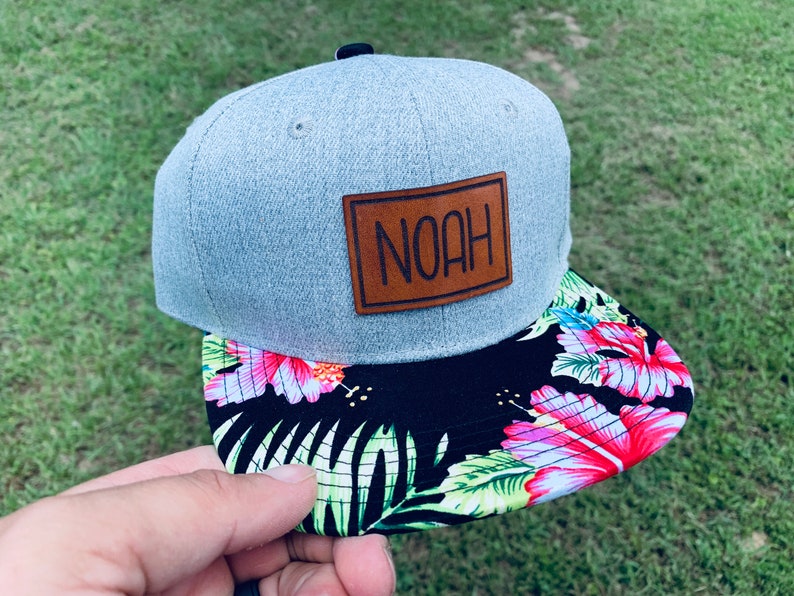May include: A gray and black baseball cap with a brown leather patch that says "NOAH" on it. The brim of the cap has a tropical print with pink flowers and green leaves.