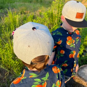 May include: Two children wearing gray baseball caps with black brims. The caps have a brown leather patch on the back. Both children are wearing blue shirts with a white and orange floral pattern.