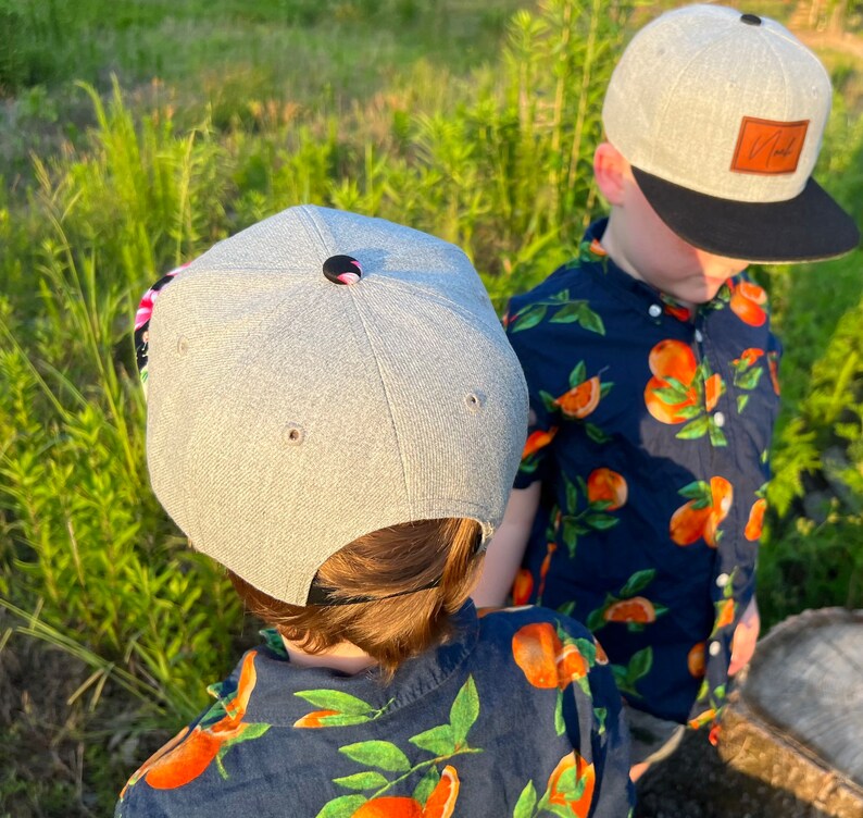 May include: Two children wearing gray baseball caps with black brims. The caps have a brown leather patch on the back. Both children are wearing blue shirts with a white and orange floral pattern.