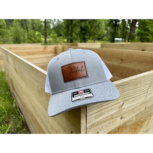 May include: A gray trucker hat with a white mesh back and a brown leather patch. The patch features the name "Judah" in a cursive font. The hat is a Richardson 112 model.