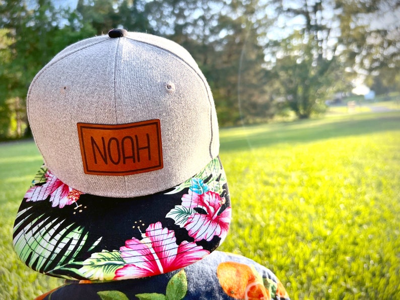May include: A gray and black baseball cap with a brown leather patch that says "NOAH" on it. The cap has a floral print on the underside of the brim.
