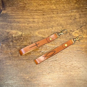 May include: Two brown leather keychains with metal key rings. One keychain has a cross design and the other has a fish design.