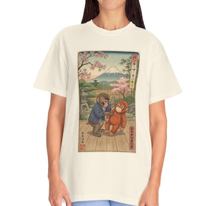 以下が含まれることがあります： クリーム色のTシャツ。日本の風景にインスパイアされたイラストが特徴です。アートワークは、背景に富士山を配した風光明媚な風景の中に2匹の猿を描いています。デザインには桜と日本語のテキストが含まれています。