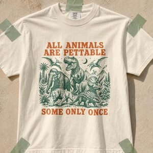 May include: An off-white short-sleeve t-shirt featuring a green graphic illustration of various prehistoric animals including a T-Rex, mammoth, and triceratops. The text reads ALL ANIMALS ARE PETTABLE SOME ONLY ONCE in orange block lettering.