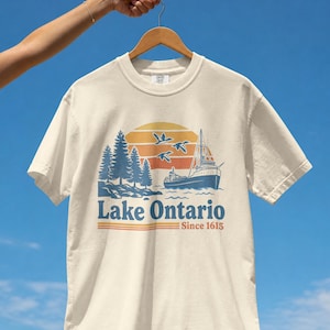 May include: A cream-colored short-sleeve t-shirt featuring a graphic of a ship on water, pine trees, and three flying birds against a sunset. The text reads Lake Ontario and Since 1615, with horizontal lines underneath the text in orange and blue.