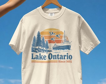 Può includere: Una t-shirt a maniche corte color crema con una grafica raffigurante una nave sull'acqua, pini e tre uccelli in volo davanti a un tramonto. Il testo riporta Lake Ontario e Since 1615, con linee orizzontali arancioni e blu sotto la scritta.