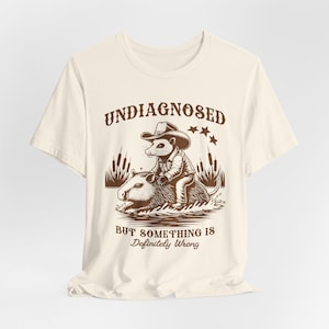 Op de afbeelding: Een beige T-shirt met een bruine afbeelding van een cartoon-opossum dat een capibara berijdt. De tekst "Undiagnosed but something is definitely wrong" staat op het shirt gedrukt.
