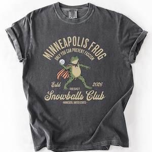 May include: Dark gray t-shirt with a graphic of a frog wearing a cape and holding a snowball. Text reads "Minneapolis Frog" and "Only You Can Prevent Fascism." Additional text includes "Snowballs Club" and "Minnesota, United States."