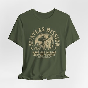 May include: Olive green t-shirt featuring a vintage-style graphic. The design includes an alien in a cowboy hat, a desert scene, and the text "31/ATLAS MISSION MARS WAS LOOKING BETTER ANYWAY!" in a circular layout. The shirt also has the text "IN NOVEMBER 2025 THE SHIP LEFT PLANET EARTH".