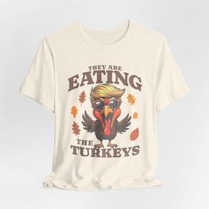Puede incluir: Una camiseta beige con un estampado gráfico de un pavo que lleva gafas de sol y una peluca rubia. El texto "They are eating the turkeys" está impreso en la camiseta.