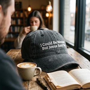 以下が含まれることがあります： 「I Could Be Meaner But Jesus Said No」の刺繍が入った黒い野球帽。帽子は、コーヒーカップと開いた本が置かれた木製のテーブルの上にあります。背景にはコーヒーを飲んでいる人がいます。