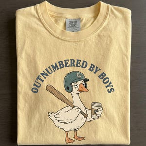 May include: A light yellow t-shirt with the text "OUTNUMBERED BY BOYS" in navy blue. The shirt features a cartoon goose wearing a baseball helmet and holding a bat and coffee cup. The shirt is folded.