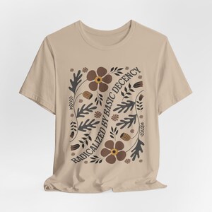 Pode incluir: Uma camiseta bege com um design floral e o texto "Radicalized by Basic Decency" em preto.