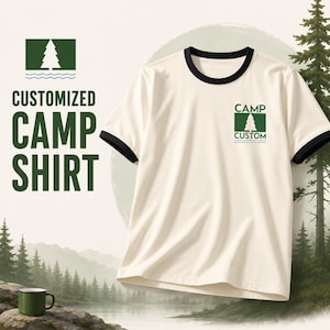 May include: A cream-colored ringer t-shirt with black trim on the collar and sleeves. The chest features a green rectangular graphic with a white pine tree icon, wavy lines, and the text "CAMP CUSTOM" printed in a dark green sans-serif font.
