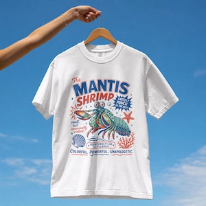 Pode incluir: Uma t-shirt branca de manga curta com um gráfico de um camarão-mantis com o texto "The MANTIS SHRIMP", "FASTEST PUNCH IN THE REEF", "small but unreasonably violent", "ODONTODACTYLUS SCYLLARUS" e "COLORFUL. POWERFUL. UNAPOLOGETIC."