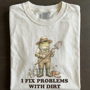 Puede incluir: Camiseta color crema con una rana de dibujos animados vestida de jardinero, sosteniendo una pala. La rana lleva sombrero, peto y botas. El texto en la parte inferior dice "I FIX PROBLEMS WITH DIRT". Diseño temático de jardinería.