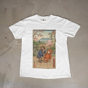 May include: White t-shirt featuring a Japanese-inspired illustration of two monkeys. One monkey is wearing a blue jacket and the other is orange. The background includes a mountain, cherry blossoms, and Japanese text.