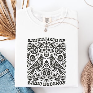 Op de afbeelding: Wit sweatshirt met een zwart grafisch ontwerp met een schedel, twee kraaien en bloemmotieven. De tekst "RADICALIZED BY BASIC DECENCY" staat eronder gedrukt. Een zilveren ketting met een hanger ligt op het shirt.