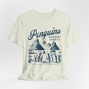 Könnte beinhalten: Ein weißes T-Shirt mit einer blauen Grafik von Pinguinen vor einer Bergkulisse. Der Text "Penguins Against Tariffs" befindet sich über den Pinguinen und "Heard Island and McDonald Islands" darunter.