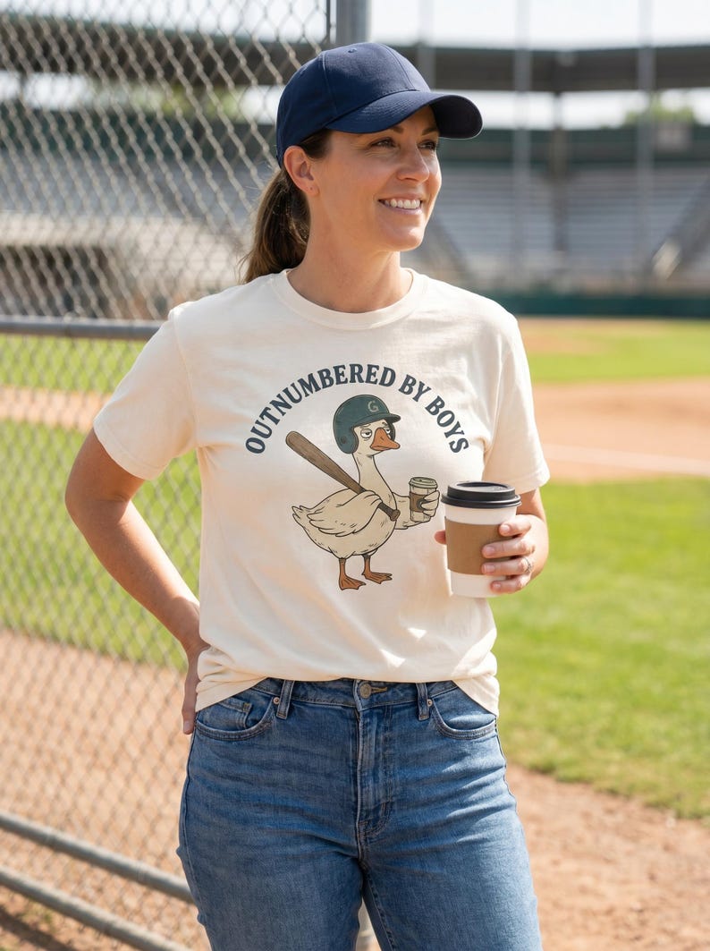 May include: Cream-colored t-shirt with the text "OUTNUMBERED BY BOYS" and a cartoon goose wearing a baseball helmet and holding a bat and coffee cup. The person is wearing a navy blue baseball cap and blue jeans.