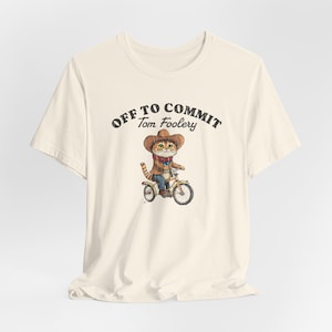 Könnte beinhalten: Cremefarbenes T-Shirt mit einer Cartoon-Katze, die einen Cowboyhut trägt und Fahrrad fährt. Der Text "OFF TO COMMIT Tom Foolery" ist über der Katzenillustration in einer verblassten Schrift gedruckt.