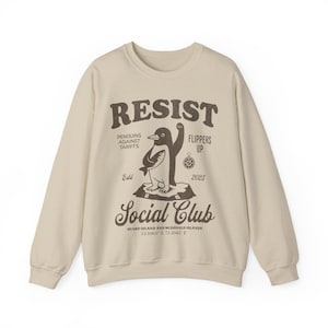 Könnte beinhalten: Ein beiger Pullover mit einem braunen Grafikdruck eines Pinguins, der einen Fisch hält. Die Grafik enthält den Text "Resist", "Penguins Against Tariffs", "Flippers Up", "Social Club", "Heard Island and McDonald Islands" und "53.1060 5, 73.5042 E".