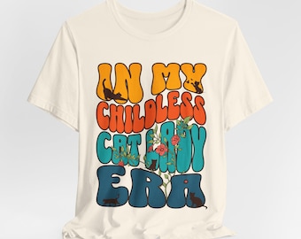 Op de afbeelding: Een wit T-shirt met een retro-stijl grafische afbeelding met de tekst "In My Childless Cat Lady Era" in kleurrijke, golvende letters. Er zijn kleine zwarte katten-silhouetten verspreid rond de tekst.