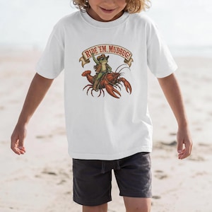 Può includere: T-shirt bianca con un cartone animato di una rana con un cappello da cowboy che cavalca un gambero rosso. Il testo "Ride 'em, Mudbug!" è visualizzato su uno striscione. Abbigliamento casual.