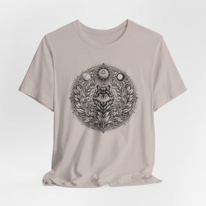 May include: A light beige t-shirt featuring a detailed black and white wolf design. The design is circular, with the wolf at the center, surrounded by leaves, moons, and geometric shapes. The shirt is made of a soft material.