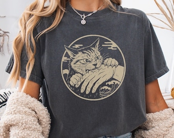 May include: Dark grey t-shirt with a circular graphic of a cat biting a hand. The cat is beige with black stripes, and the hand is beige. The design includes wave and cloud elements. The shirt is made of a soft material.