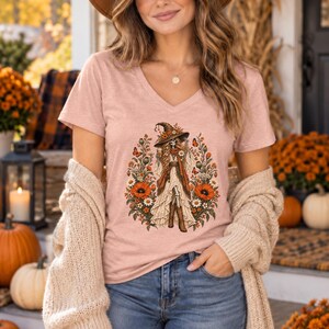 Può includere: Una t-shirt con scollo a V color rosa antico con una grafica centrale raffigurante uno scheletro che indossa un cappello da strega, una gonna lunga e stivali, circondato da una composizione floreale di papaveri arancioni, margherite e farfalle.