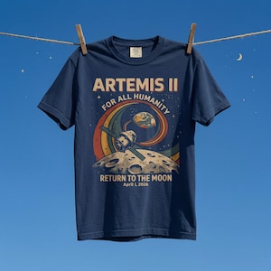 May include: Navy blue t-shirt featuring a retro graphic design with the text "ARTEMIS II FOR ALL HUMANITY" and "RETURN TO THE MOON April 1, 2026". The design includes a space station, Earth, and the moon. The shirt is hanging on a clothesline.