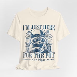 Puede incluir: Una camiseta beige con un gráfico azul de un mapache con gafas de sol y una gorra de béisbol. El mapache está sosteniendo cartas de juego y fichas. El texto "I'm just here for the pot" está impreso en la camiseta, junto con el texto "Las Vegas".