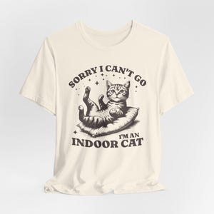 以下が含まれることがあります： 猫が枕の上に横たわっているグラフィックが特徴のクリーム色のTシャツ。「SORRY I CAN'T GO」と「I'M AN INDOOR CAT」の文字が猫の上と下にヴィンテージフォントでプリントされています。このシャツは柔らかい素材で作られています。