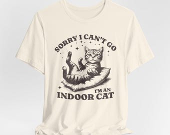 May include: Cream-colored t-shirt featuring a graphic of a cat lounging on a pillow. The cat is surrounded by the text "SORRY I CAN'T GO" and "I'M AN INDOOR CAT" in a vintage style font. The shirt is made of a soft material.