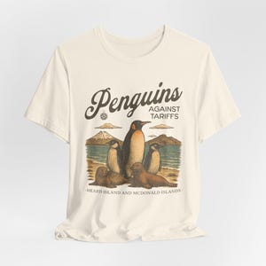 Könnte beinhalten: Ein beiges T-Shirt mit einem Aufdruck von Pinguinen gegen Zölle. Der Aufdruck zeigt eine Gruppe von Pinguinen, die an einem Strand stehen, mit dem Text "Penguins Against Tariffs" und "Heard Island and McDonald Islands".