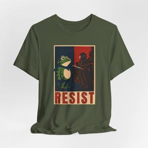 May include: Olive green t-shirt with a graphic of a frog wearing a scarf, set against a blue and red backdrop. The word "RESIST" is printed in red block letters below the frog.