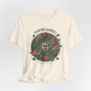 May include: A cream-colored t-shirt with a vintage-style graphic of a planetary alignment. The graphic features a sun with a face, planets, and stars. The text "Planetary Alignment" and "January 25, 2023" is printed on the shirt.