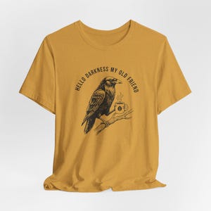 Peut inclure: T-shirt jaune moutarde avec une illustration noire d'un corbeau tenant une tasse de café sur une branche. Le texte "HELLO DARKNESS MY OLD FRIEND" est arqué au-dessus du corbeau. Le t-shirt est en matière souple.