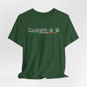 May include: A dark green t-shirt with the Irish phrase "Tiocfaidh ár lá" printed in white on the front. The phrase is accompanied by a stylized Irish flag in green, white, and orange.
