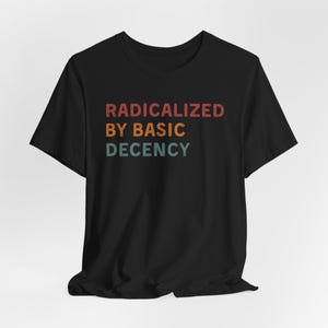 Op de afbeelding: Zwart T-shirt met de tekst "RADICALIZED BY BASIC DECENCY" in een retro lettertype met rode, oranje, groene en blauwe kleuren.
