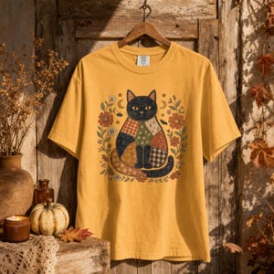 May include: A mustard yellow short-sleeve t-shirt featuring a central graphic of a cat with a patchwork-style body pattern. The cat is surrounded by a floral wreath with small stars, moons, and bats. The shirt has a crew neckline and a tag.