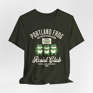Puede incluir: Camiseta verde oscuro con el texto "Portland Frog Resist Club" y un gráfico de tres ranas sosteniendo un cartel que dice "Frogs Together Strong". La camiseta también incluye el texto "Only You Can Prevent Fascism" y "Portland, Oregon 2025."